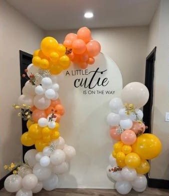 Peach, yellow and white balloon arch around a backdrop reading A little cutie is on the way