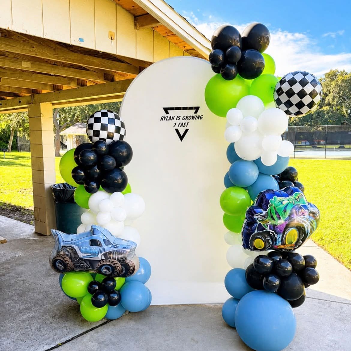 Green, black and blue monster-truck themed balloon arch around a white backdrop at a park pavilion