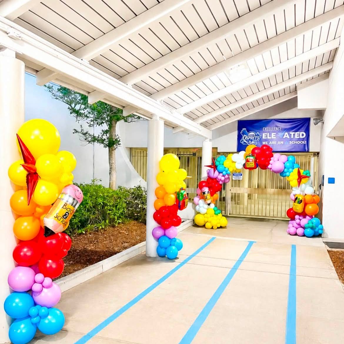 A school walkway lined with rainbow balloon columns leading to a balloon arch