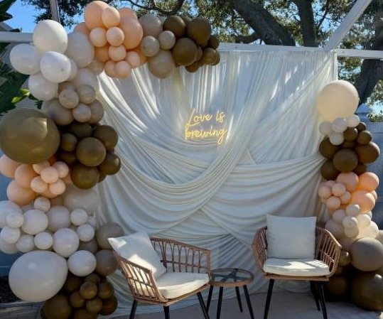 Neutral cream and olive balloon garland framing a draped backdrop with a 'Love is brewing' neon sign
